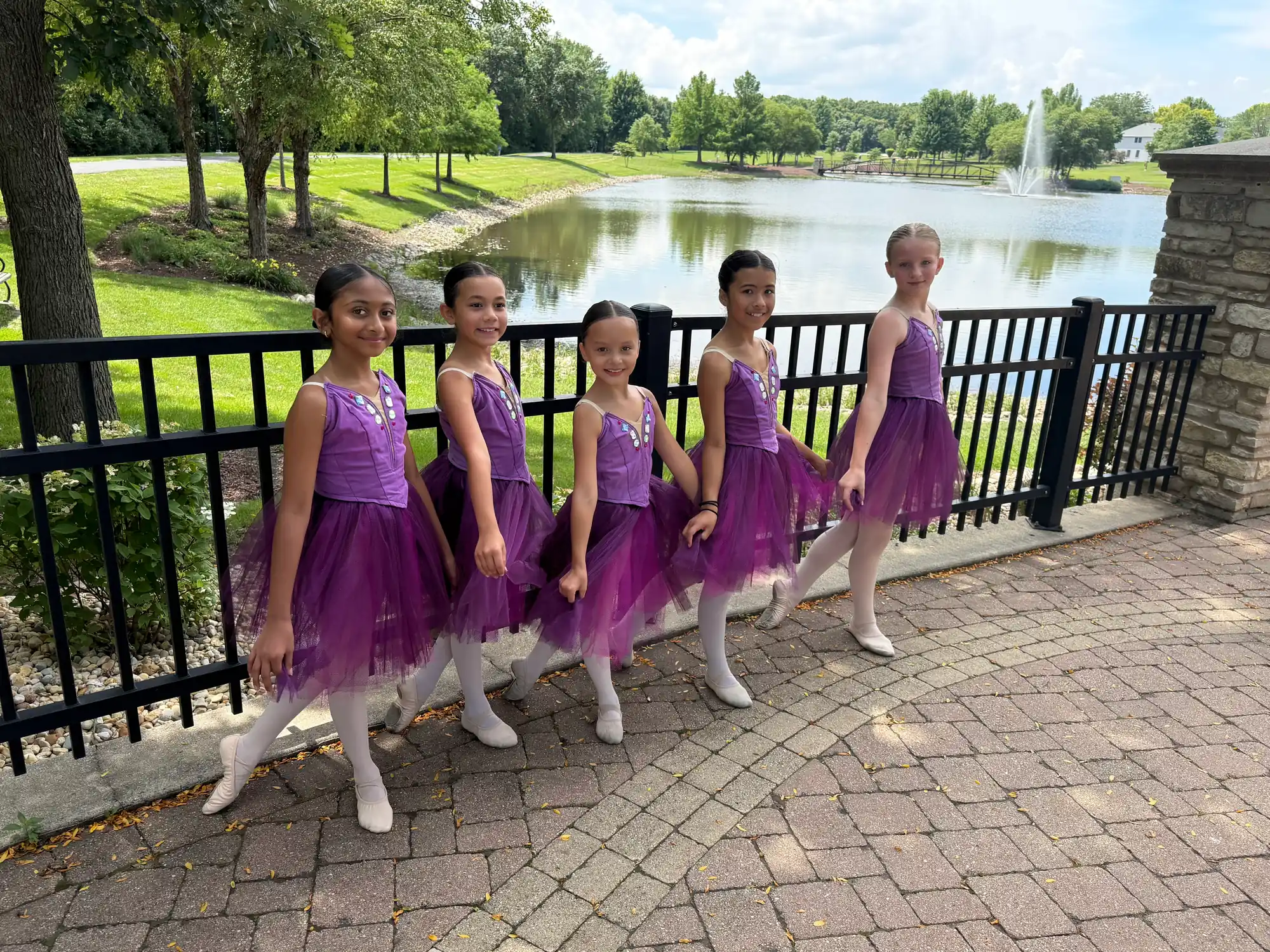 Five young dancers in purple dresses posing on a brick walkway beside a pond