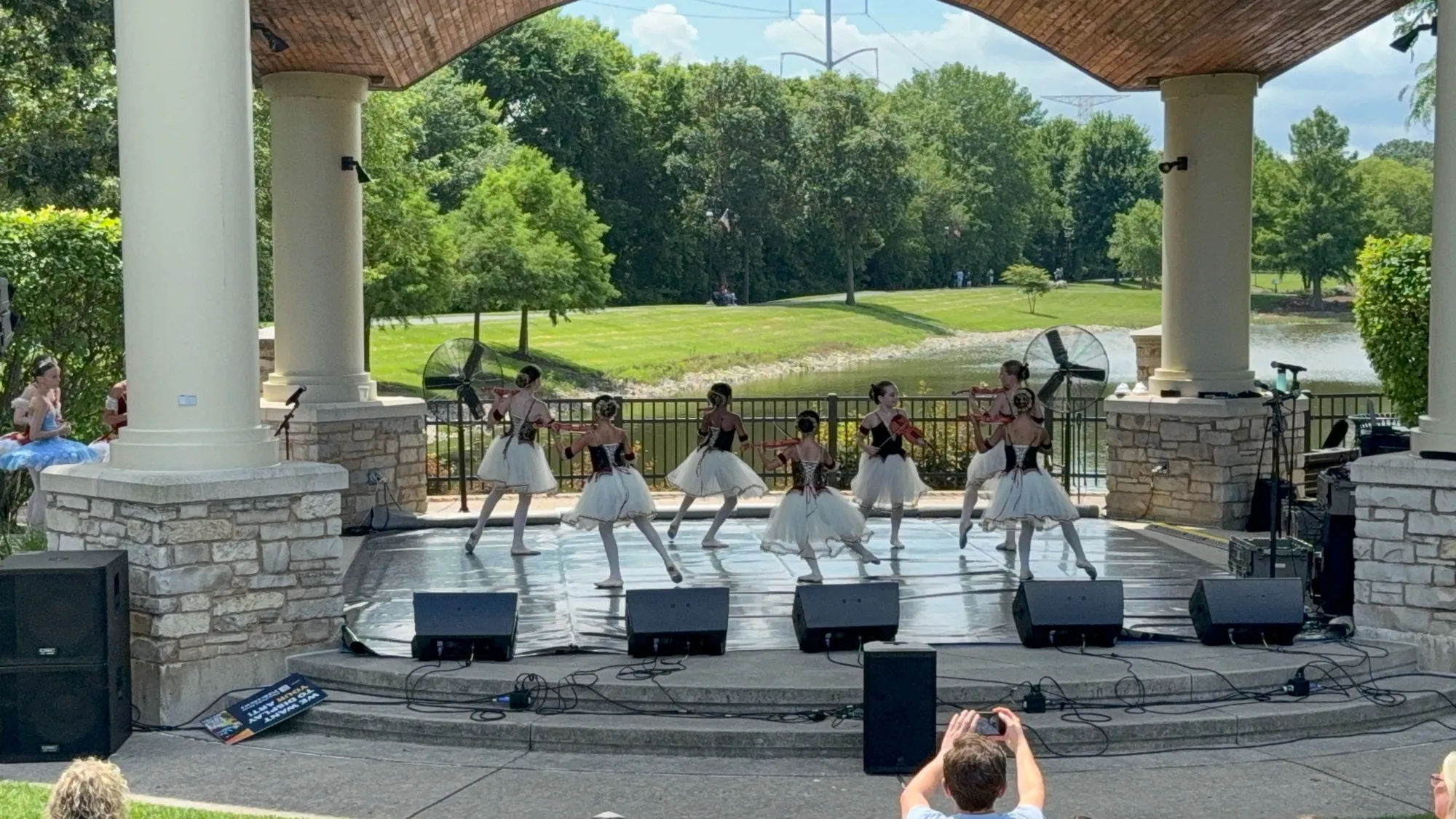 Dancers in white tutus on an outdoor pavilion stage, with trees and a lawn behind them