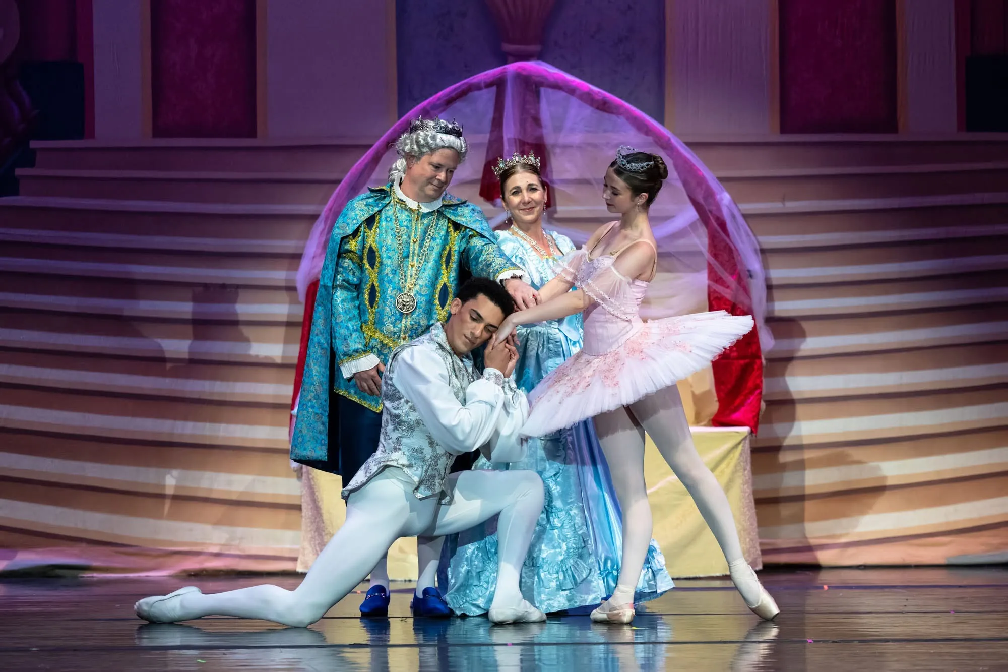 A dancer kneels to a dancer in a pink tutu, with two dancers in royal costumes behind them on stage