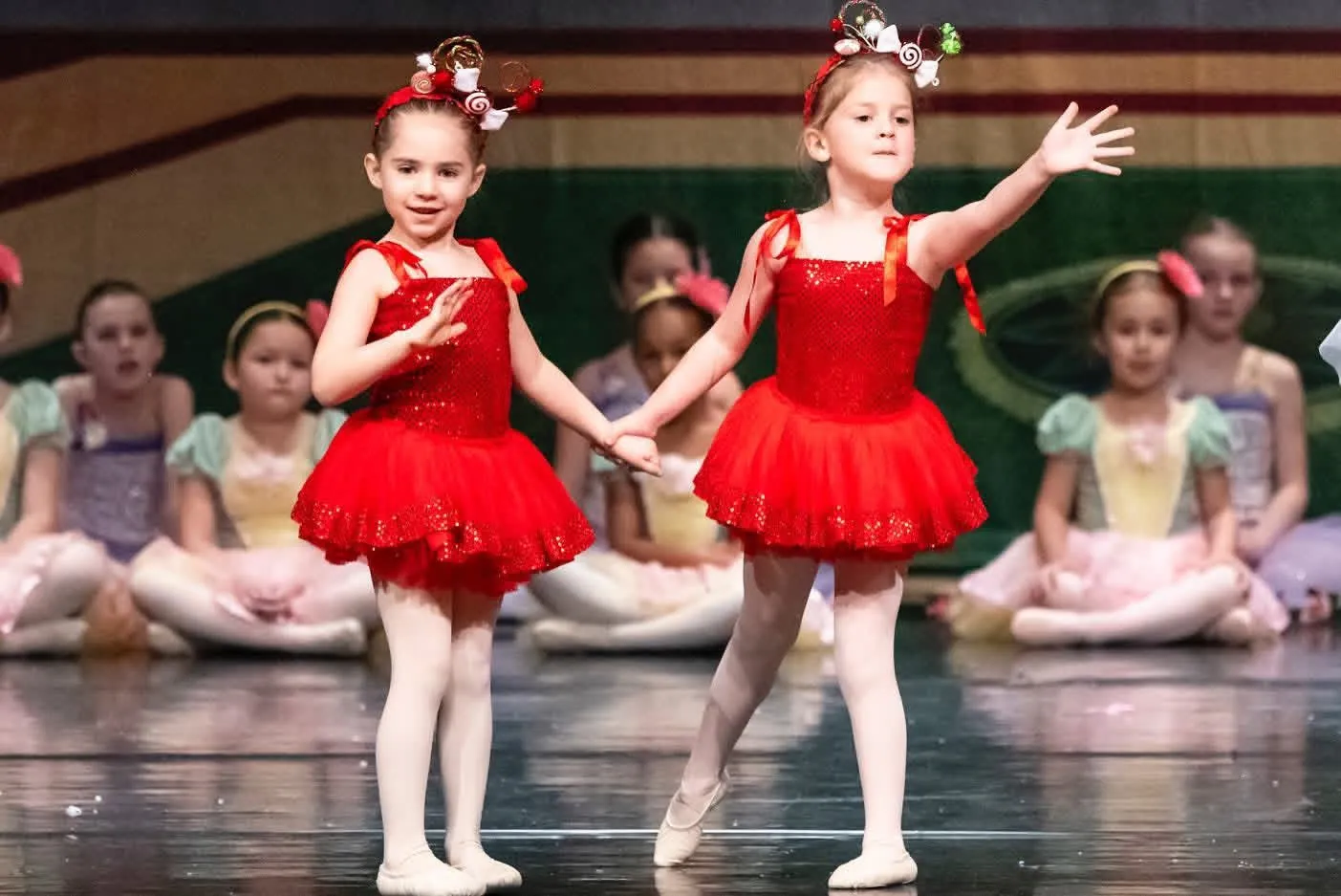 Two young dancers in red tutus and flower headpieces dancing on stage, seated dancers in pastel costumes behind them
