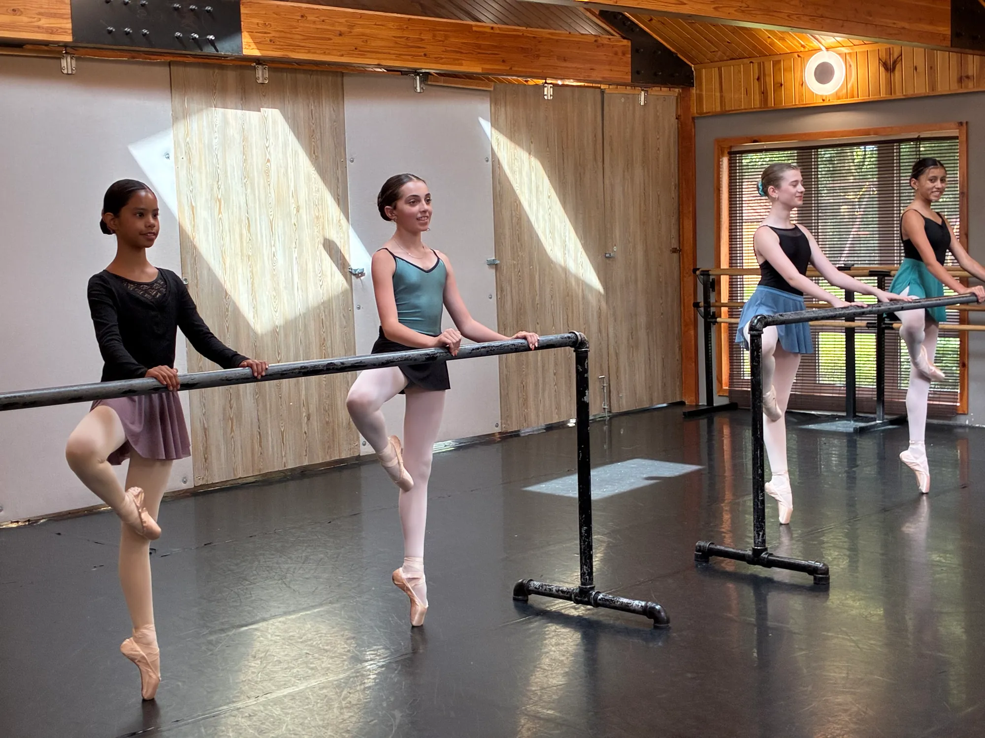 Four dancers in leotards on pointe at the barre in a sunlit studio