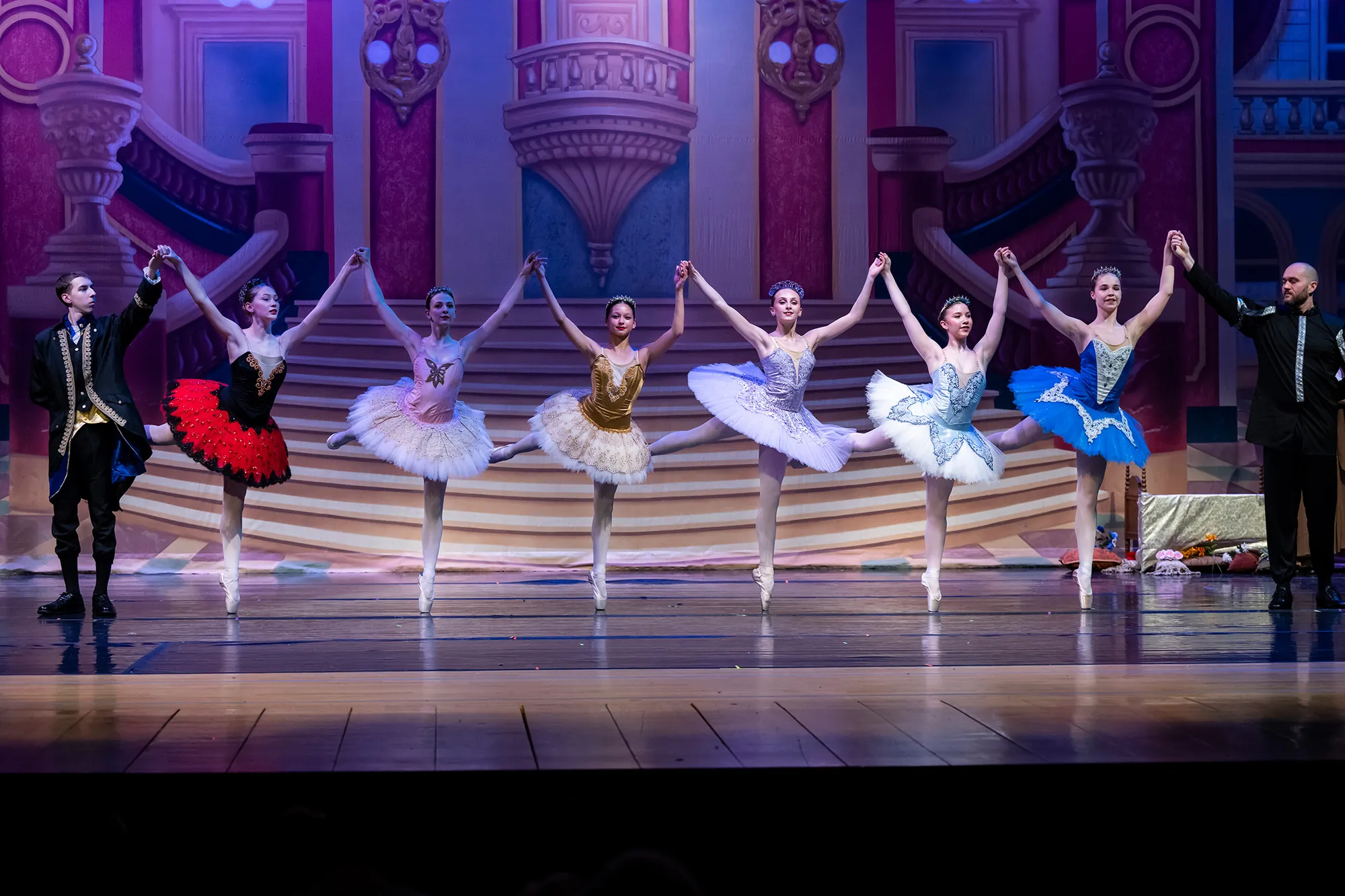 A line of dancers in tutus of different colors with arms raised on stage