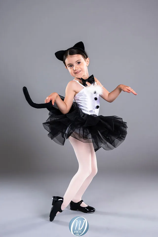 A young dancer in a black and white cat costume with ears and a tail, posing against a gray backdrop