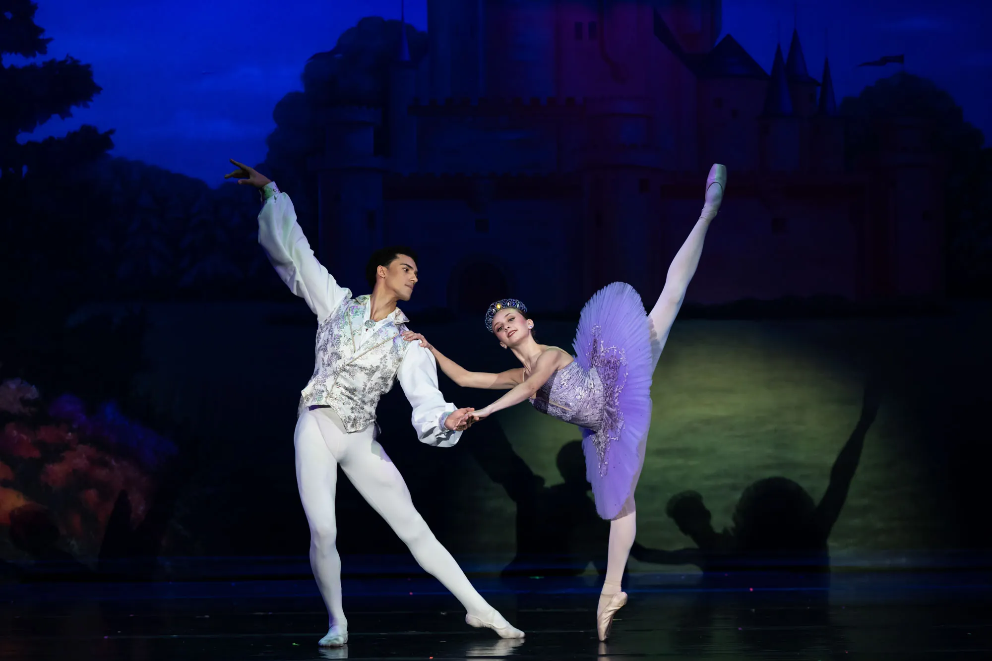 A dancer in white and a dancer in a lilac tutu in a partnered pose on a moonlit stage set