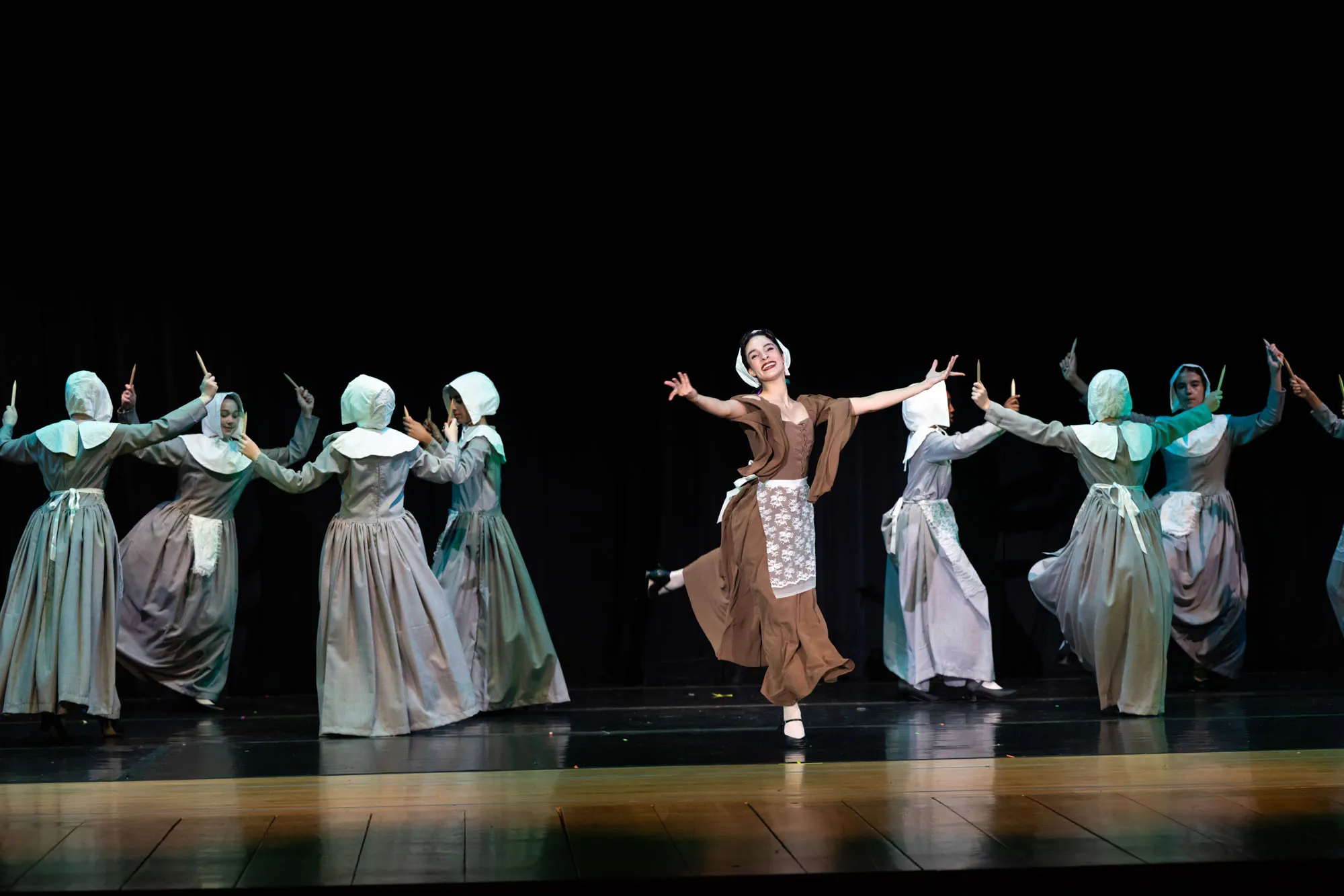 Dancers in long gray and green dresses and head scarves in a scene on a dark stage