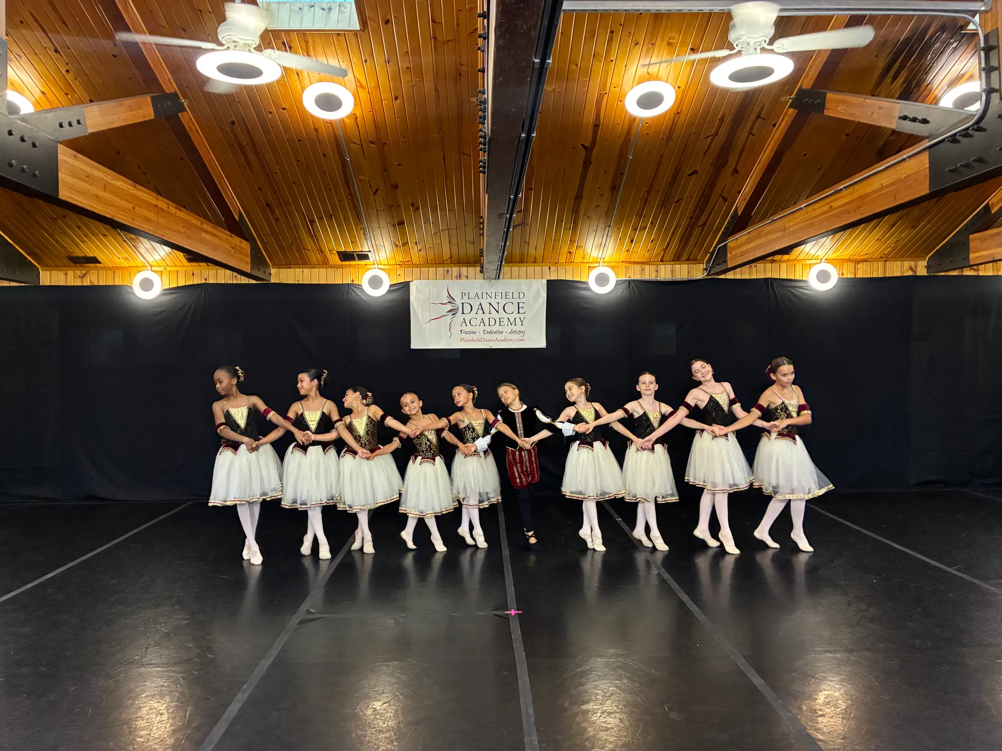 A line of dancers in white tutus on a black dance floor, with a Plainfield Dance Academy banner behind them