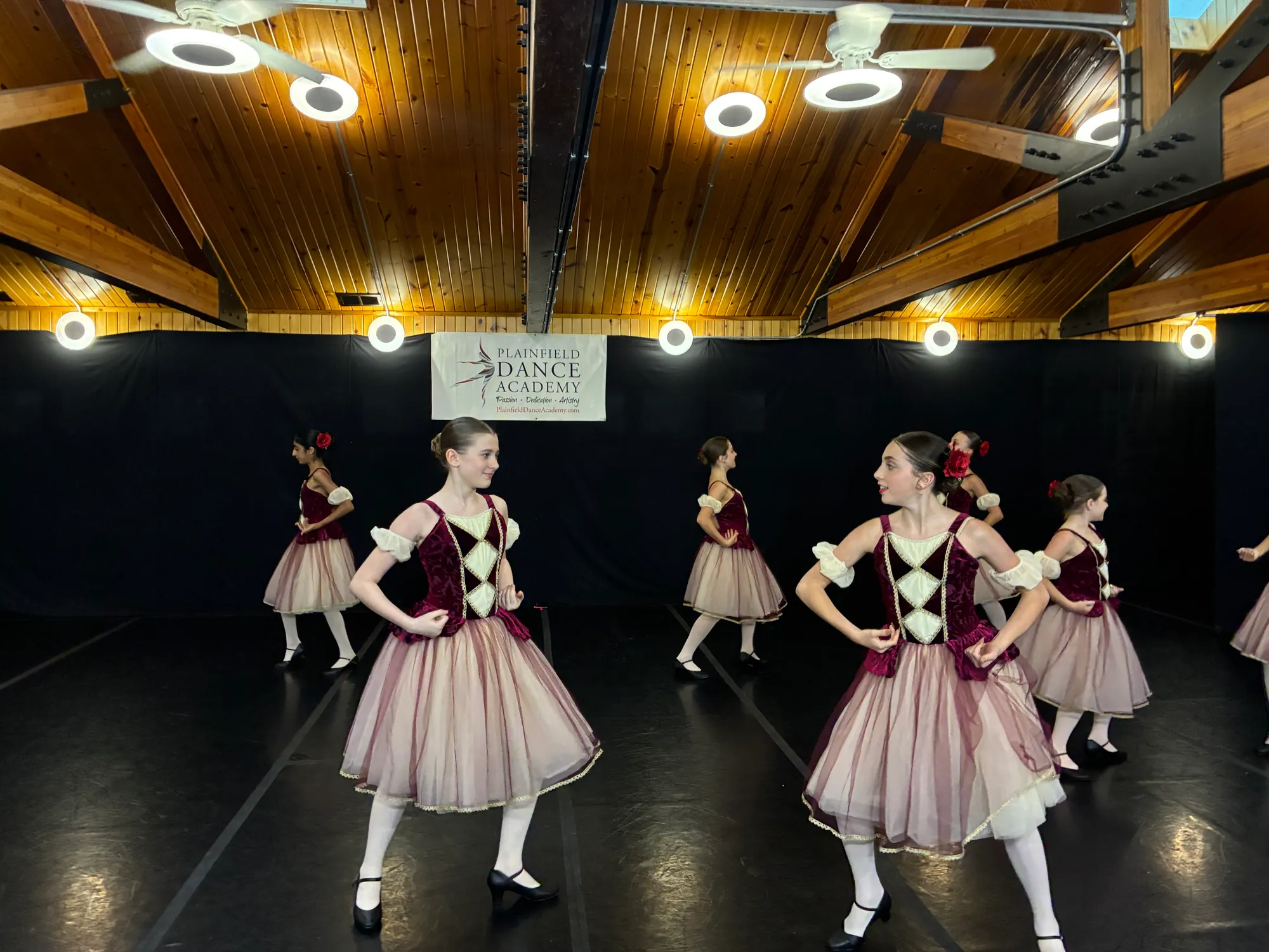 Dancers in burgundy and pink character costumes on a black dance floor, with a Plainfield Dance Academy banner behind them