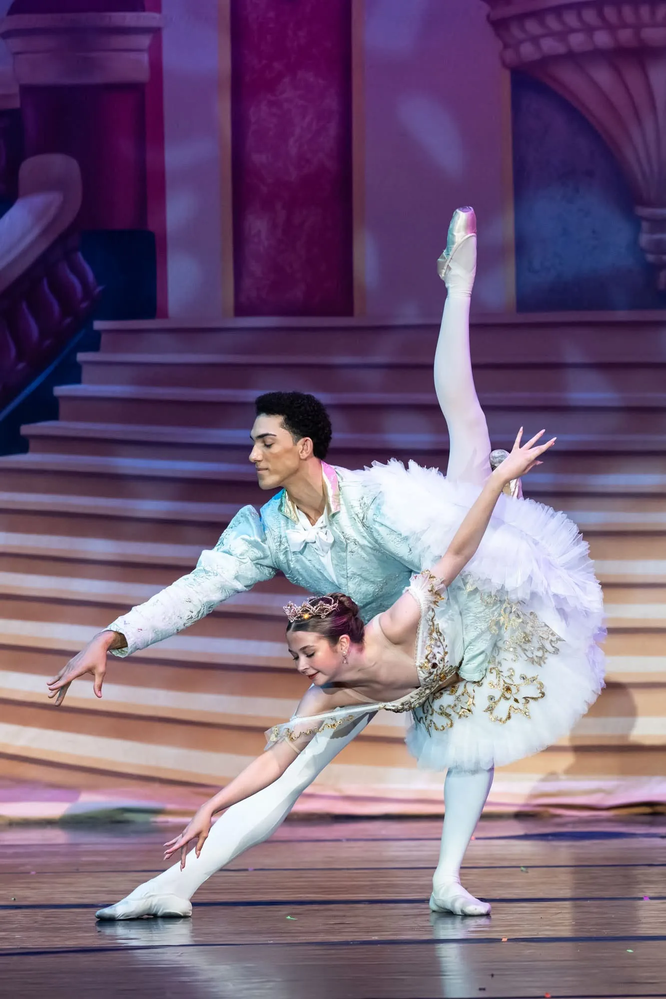 Two dancers in pale blue and white in a deep partnered pose on a stage staircase set