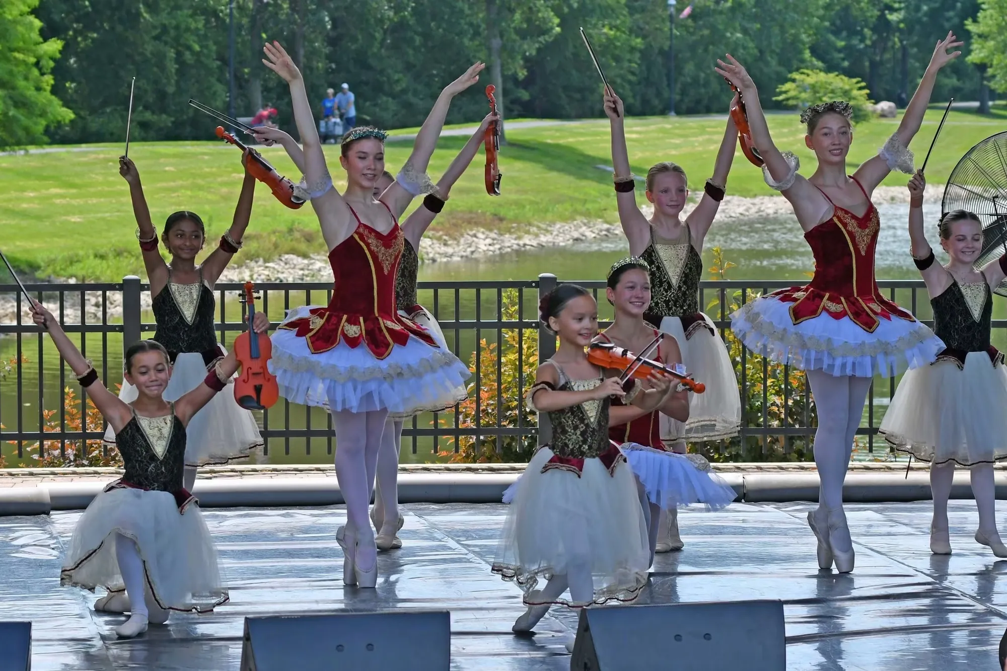 Dancers in red, white and green costumes, some holding violins overhead, on an outdoor stage