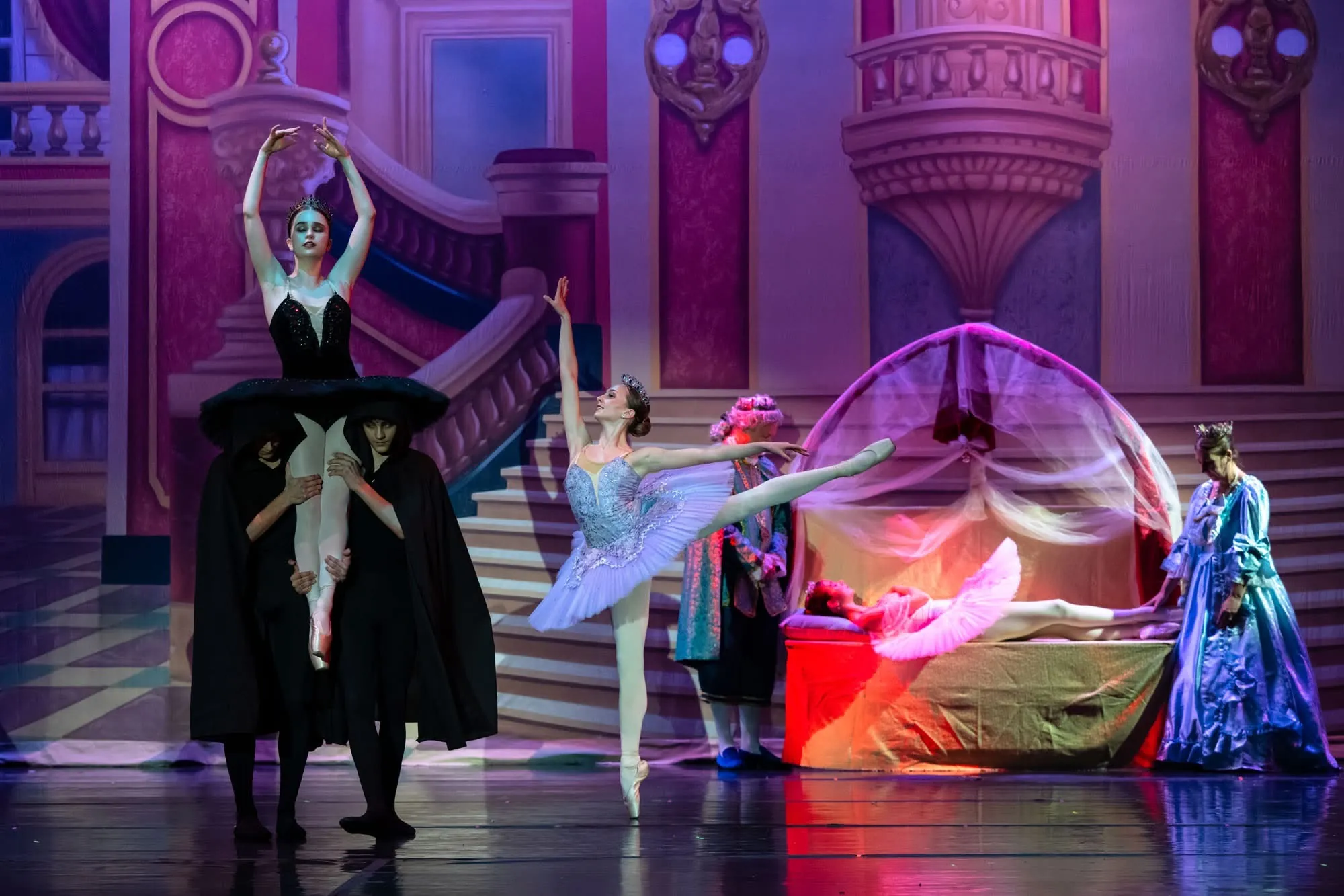 A dancer in a pale blue tutu in arabesque beside a dancer lifted overhead in a palace stage scene
