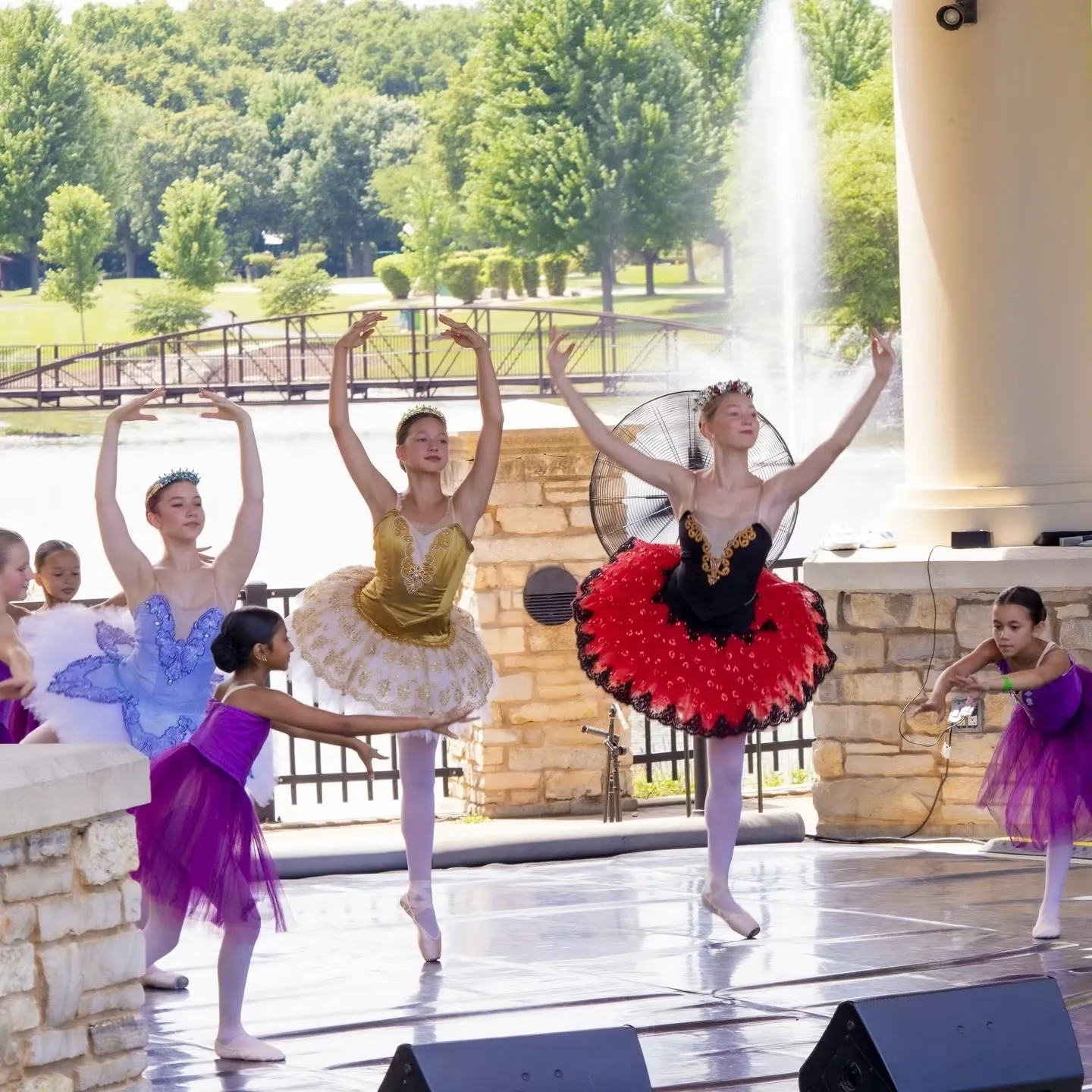 Dancers in gold, blue, red and purple tutus with arms raised on an outdoor stage beside a fountain