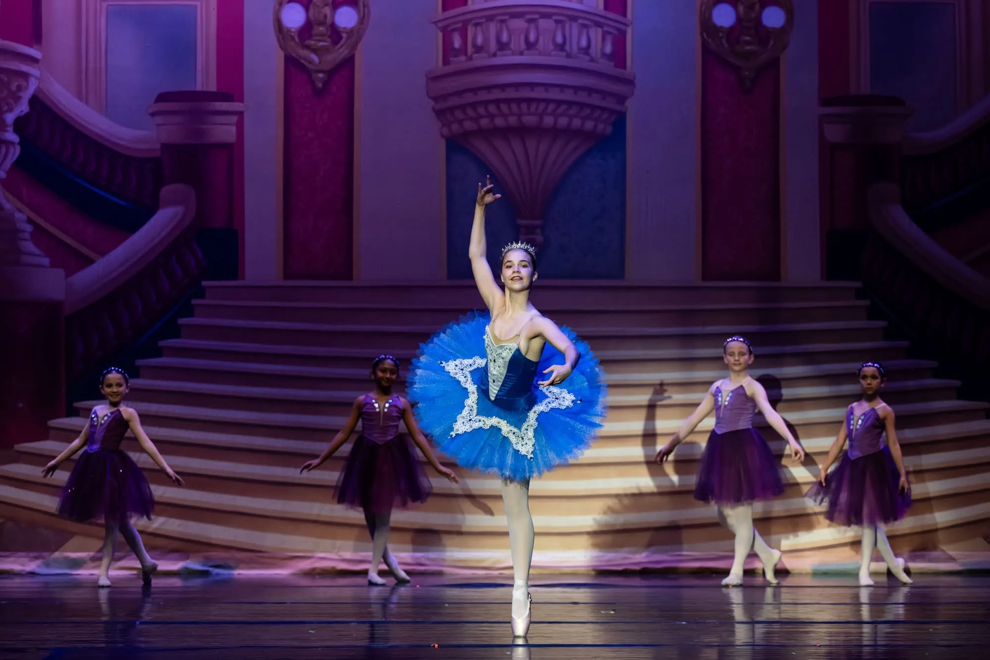 A dancer in a blue tutu on pointe with one arm raised, dancers in purple around her on a palace stage set