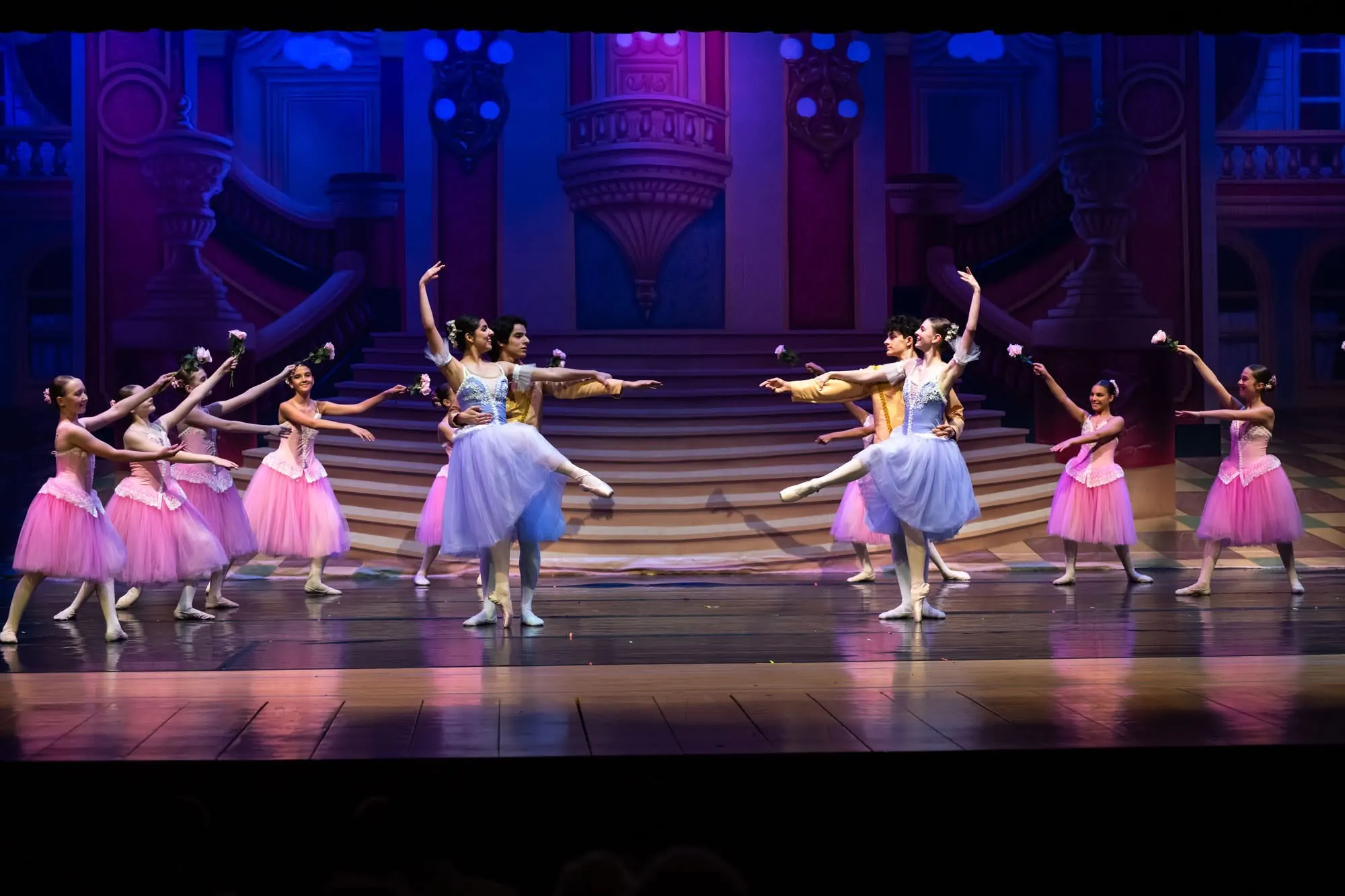 Dancers in pink and lilac costumes with arms raised on a palace stage set under blue light